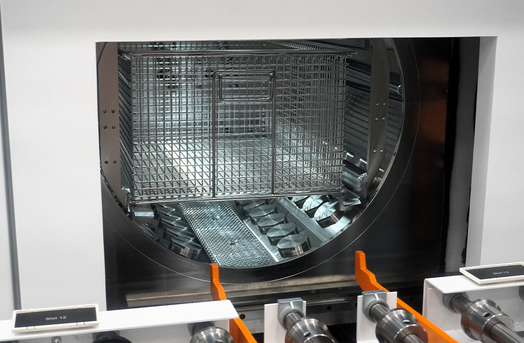De manden met werkstukken worden automatisch aangevoerd in de reinigingsmachine. Na de reiniging worden de manden via een transportband en een sluis in de cleanroom gebracht. De foto toont duidelijk de ‘hockeypuck’-modules die zorgen voor een gelijkmatige verdeling van ultrasone energie. Dampontvetter