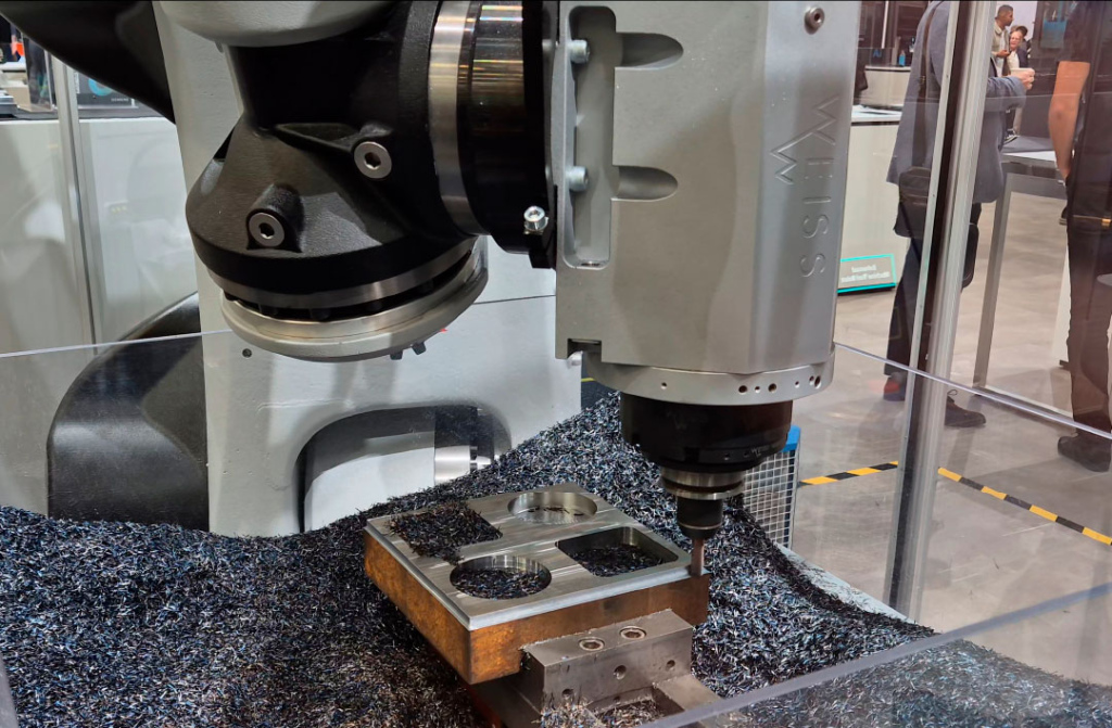 Op de foto is te zien hoe een industriële robot, aangestuurd met Siemens Sinumerik Run MyRobot, wordt ingezet voor verspanende bewerkingen in C45-constructiestaal met een treksterkte van ongeveer 570 tot 700 N/mm². De robot voert hierbij twee verschillende bewerkingsstrategieën uit. In de eerste stap wordt trochoïdaal kamerfrezen toegepast. Deze strategie is bij uitstek geschikt voor robotbewerking omdat de snijkrachten gelijkmatig worden verdeeld en piekbelastingen worden vermeden. De bewerking vindt plaats met een spiltoerental van 9.000 min⁻¹ en een voedingssnelheid van 2.000 mm/min. De snijsnelheid bedraagt 160 m/min, terwijl de radiale snedediepte is ingesteld op 0,6 mm en de axiale snedediepte op 10 mm. Deze combinatie van parameters zorgt voor een stabiele verspaning en beperkt ongewenste trillingen in de robotstructuur. Aansluitend voert de robot een bewerking uit waarbij het materiaal langs de zijkant van het werkstuk wordt gefreesd. Ook bij deze bewerking wordt gebruikgemaakt van een spiltoerental van 9.000 min⁻¹ en een voeding van 2.000 mm/min. De radiale en axiale snededieptes zijn identiek aan die van de trochoïdale bewerking, met respectievelijk 0,6 mm en 10 mm.